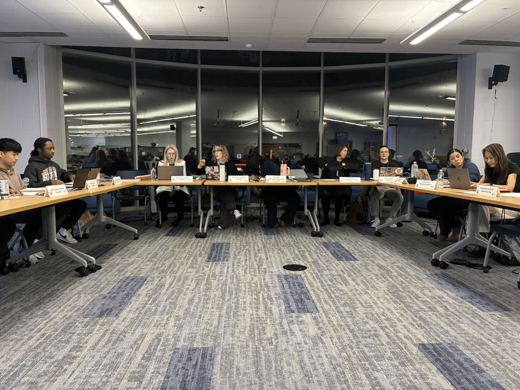 Members of Payton’s Local School Council sit around a set of tables with one table in the center and one off to each side (with these two facing each other) prior to their first meeting discussing Principal Shabazz’s contract renewal.